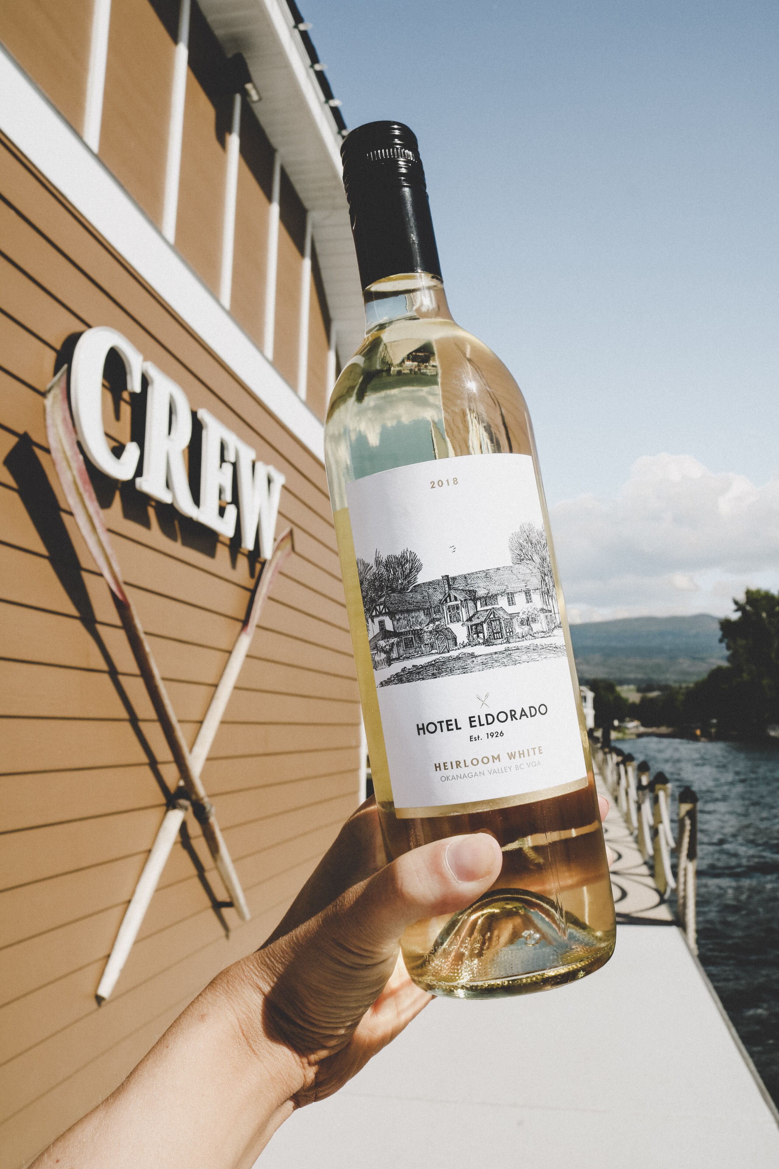 A bottle of Hotel Eldorado Heirloom White wine held by a person, with a wooden boat deck and a 'CREW' sign in the background.