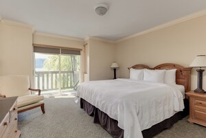 Quarto com uma cama grande, uma cadeira, uma mesa de cabeceira e janela com vista para árvores.