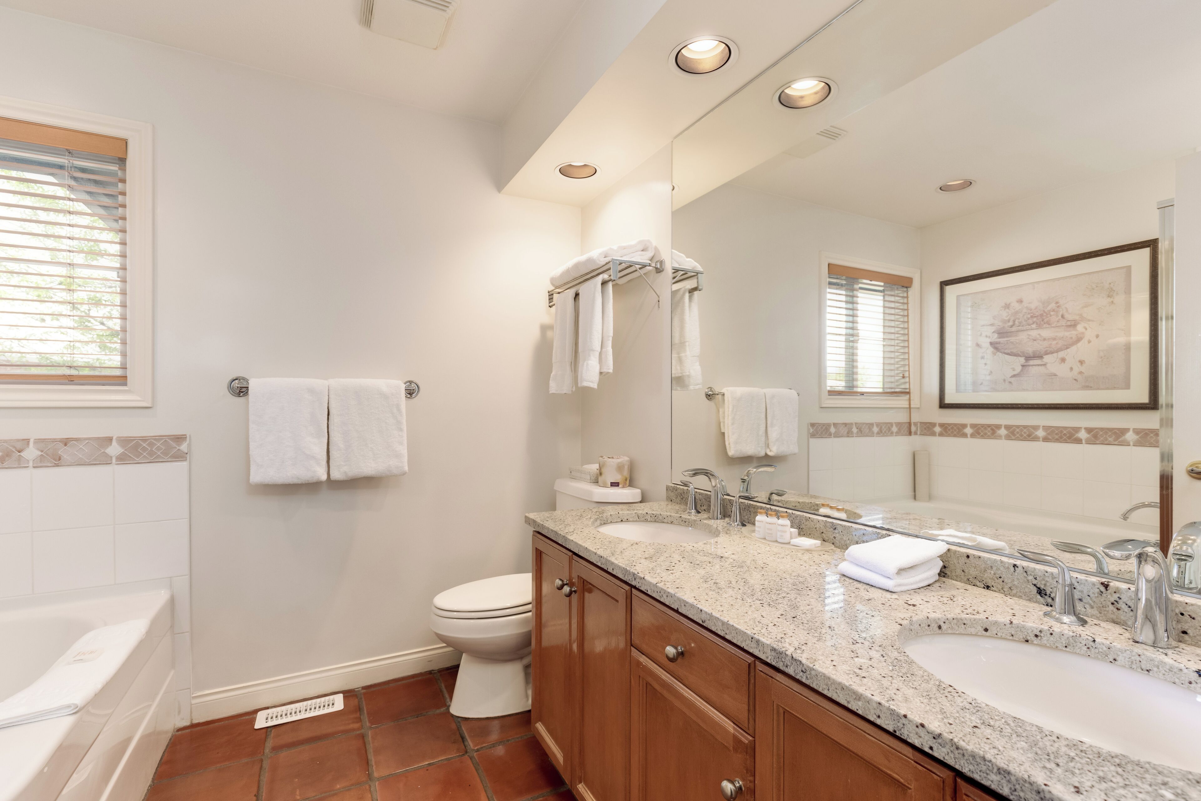 A bathroom with a double sink, toilet, and bathtub. There are towels hanging on the wall and a framed picture above the mirror.