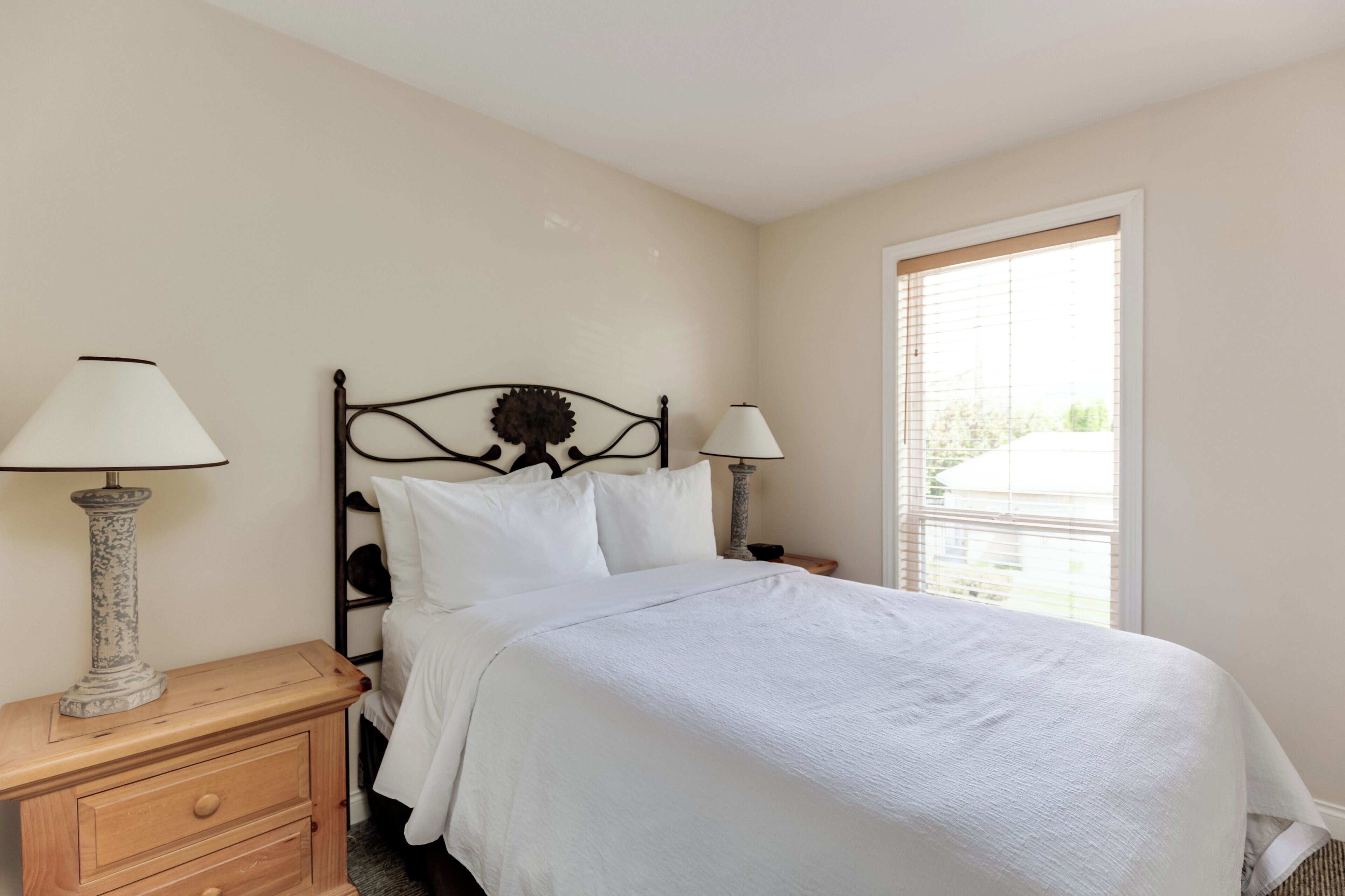 A neatly made bed with white bedding, a wooden nightstand with two lamps, and a window with blinds.