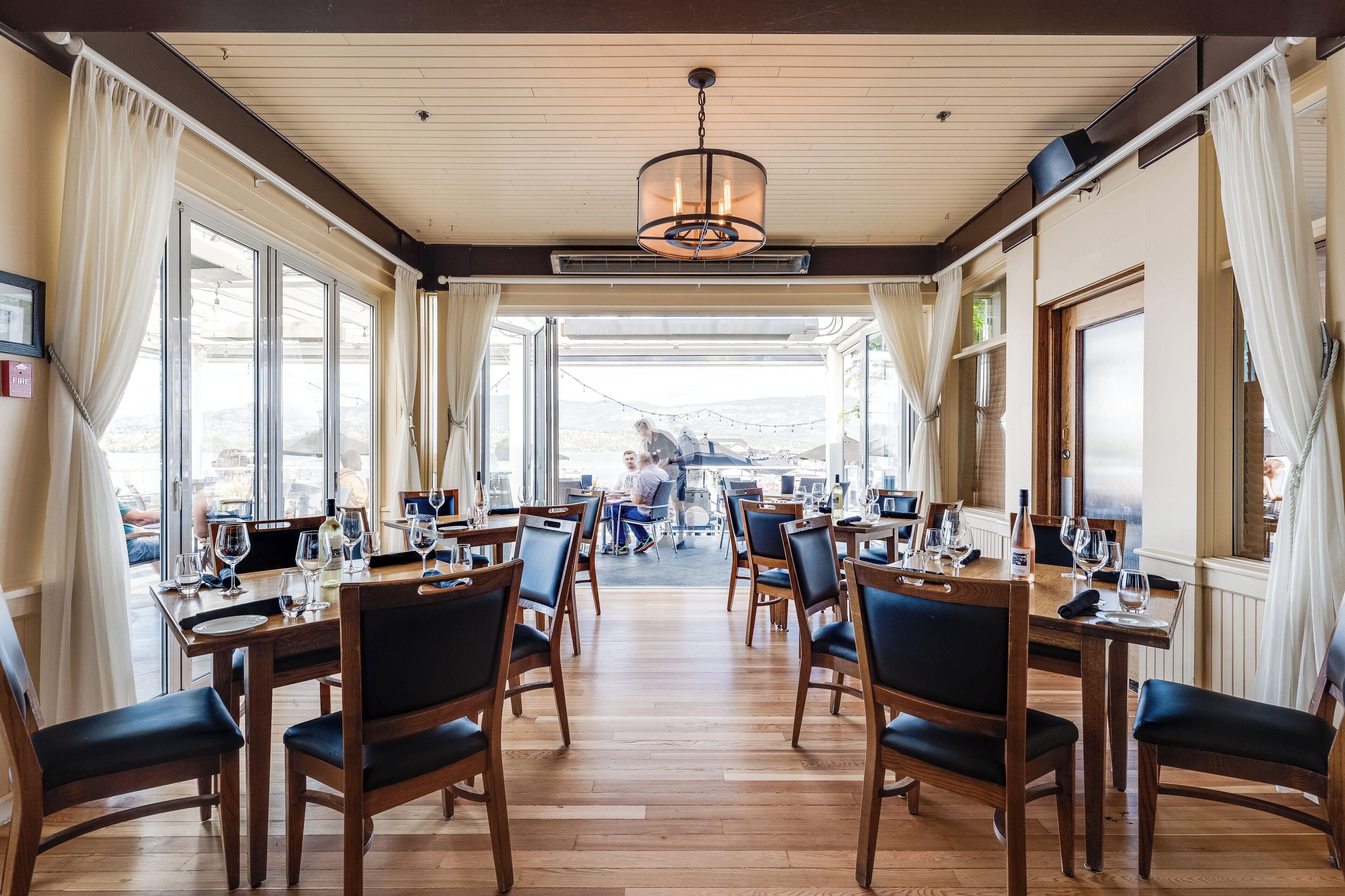 A dining area with wooden tables and chairs, large windows, and a chandelier.