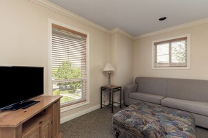 Una habitación con televisión de pantalla plana sobre un soporte de madera, un sofá gris, un otoman estampado, una mesita de noche con lámpara y una ventana con persianas.