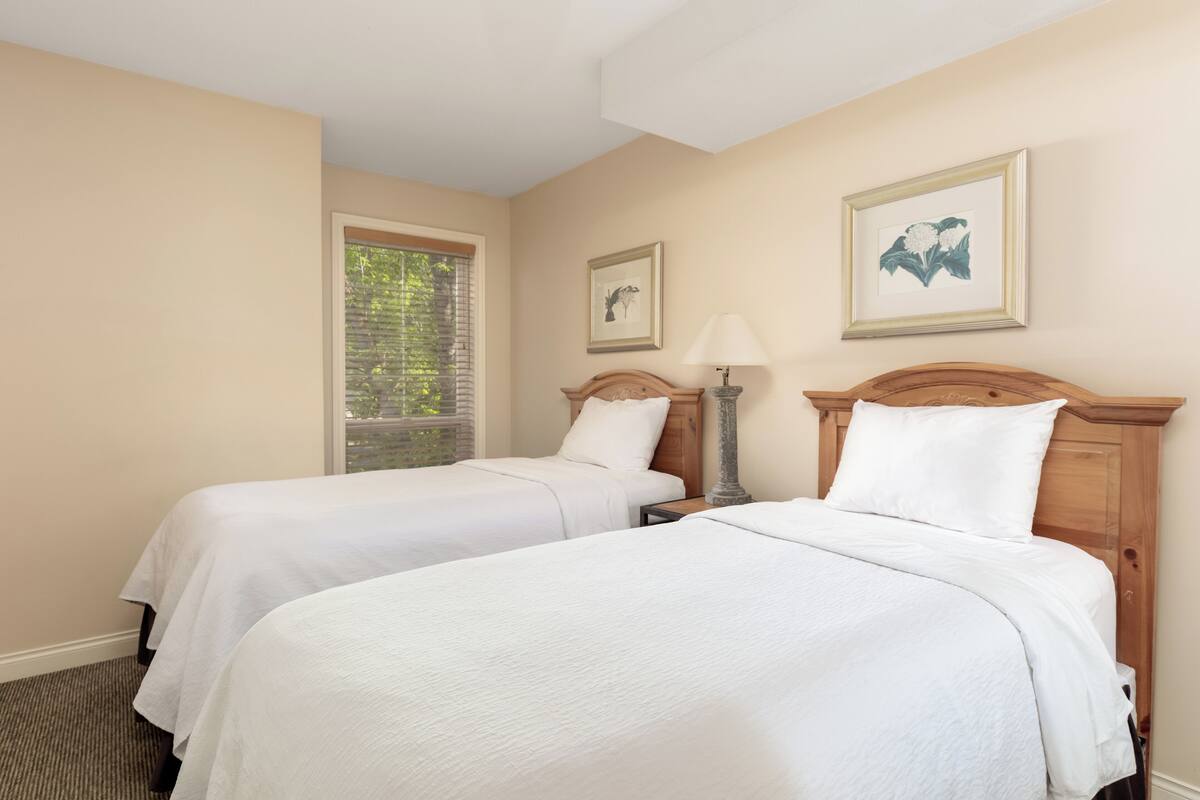 A hotel room with two single beds, a window with blinds, a bedside table with a lamp, and framed artwork on the wall.