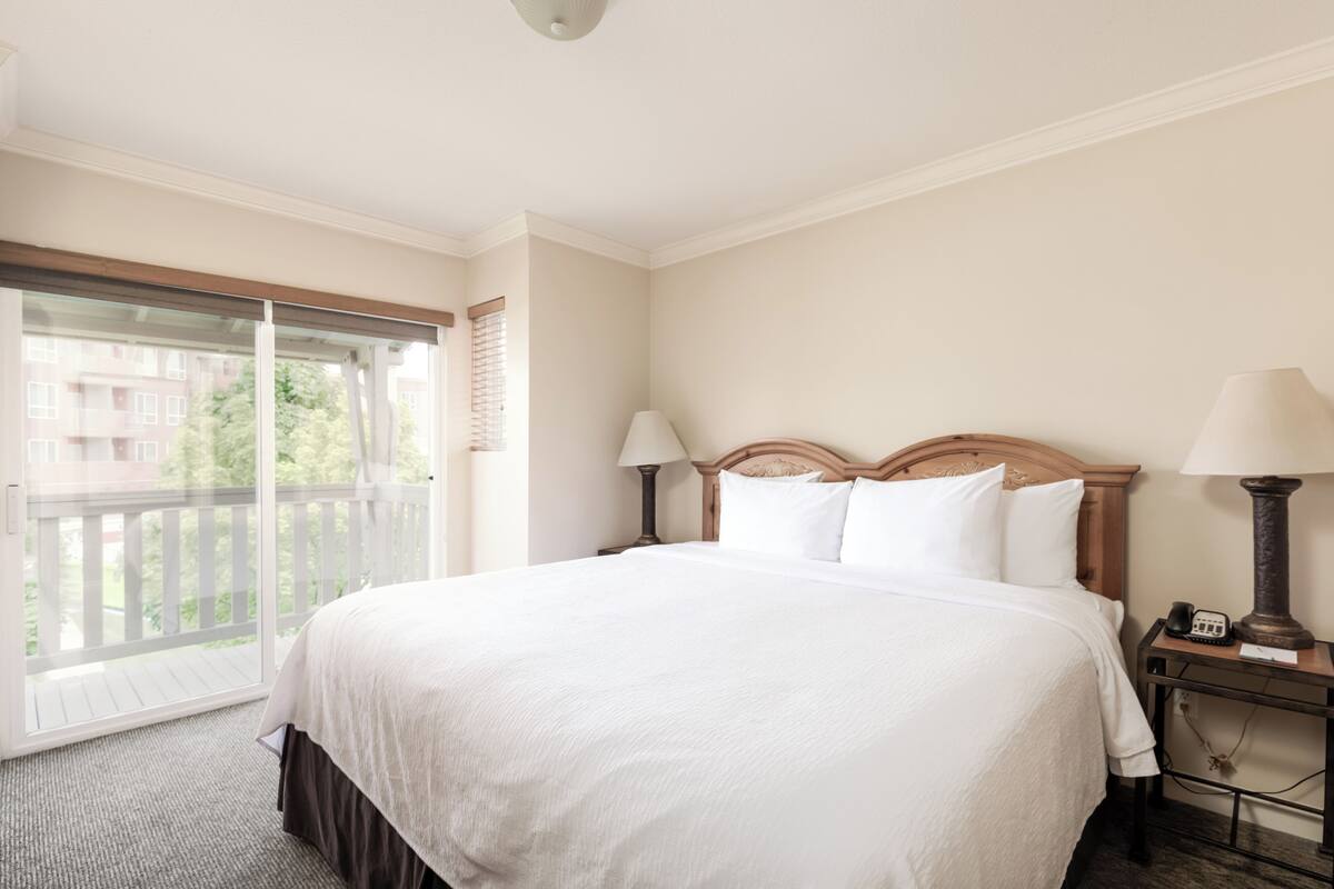 A hotel room with a large bed, two bedside tables with lamps, a sliding glass door to a balcony, and a view of buildings outside.