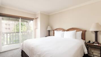 A hotel room with a large bed, two bedside tables with lamps, a sliding glass door to a balcony, and a view of buildings outside.