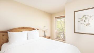 A neatly made bed with white linens in a room with a wooden headboard, a bedside lamp, and a window with blinds.