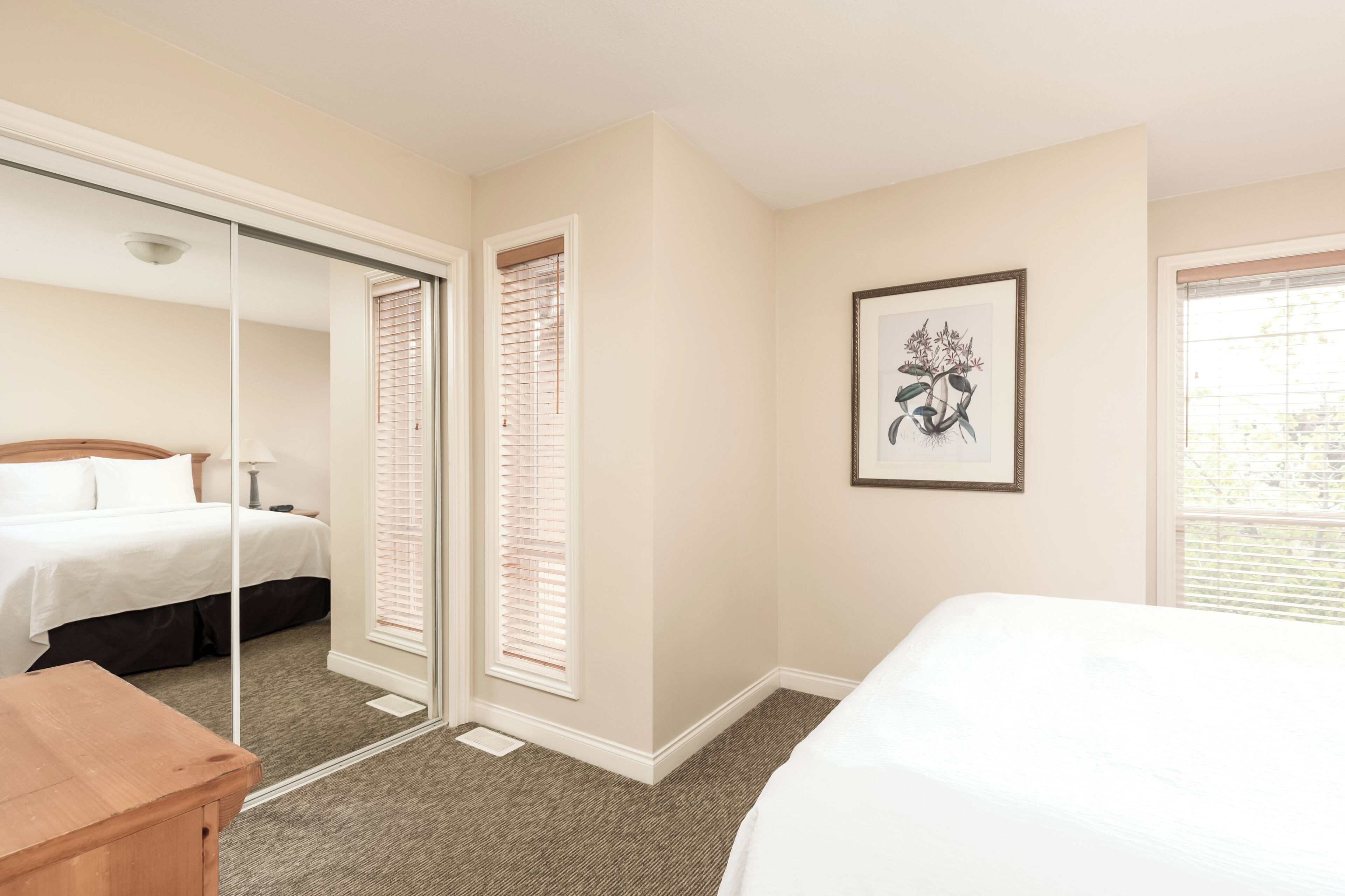 A hotel room with a bed, a wooden nightstand, a mirror, a framed artwork, and a window with blinds.