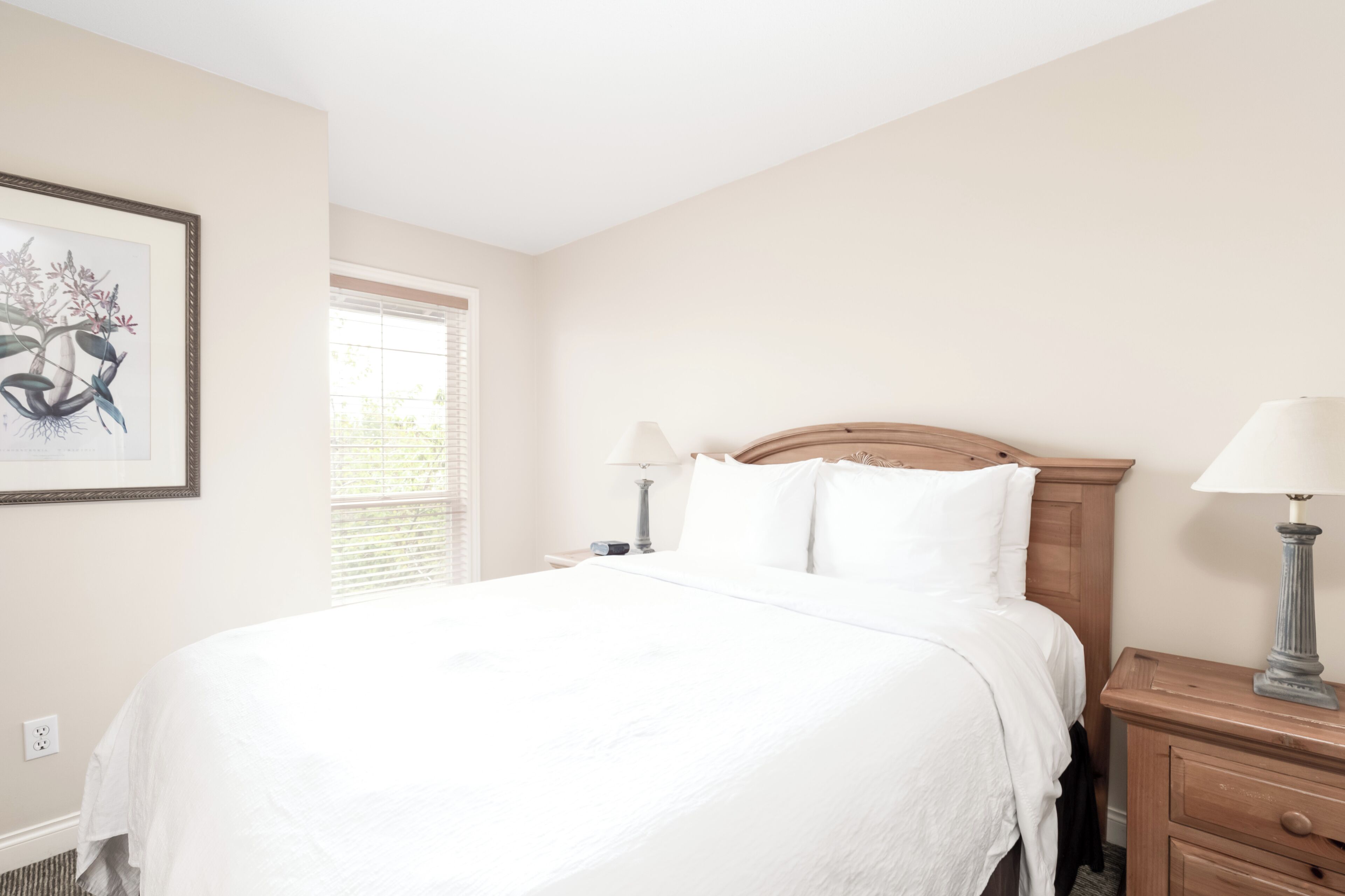 A neatly made bed with white linens, a wooden headboard, two bedside lamps, and a framed artwork on the wall.