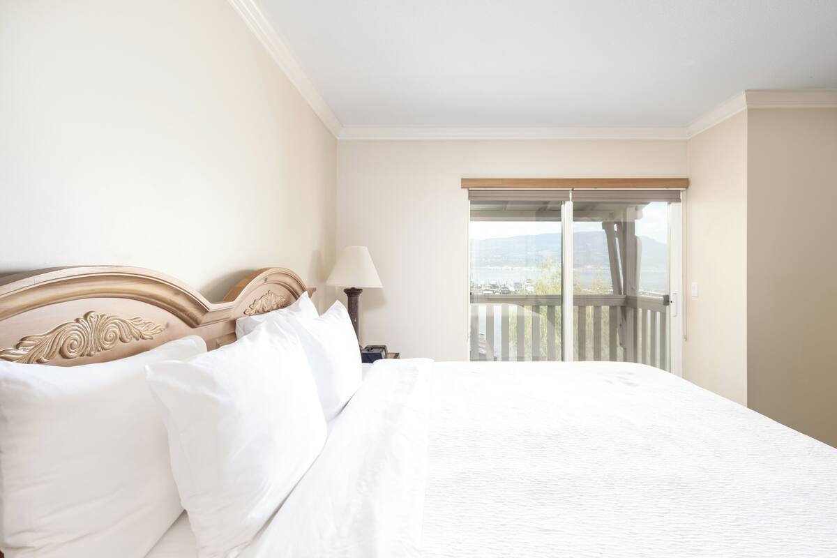 A hotel room with a large bed, a nightstand, a lamp, and a view of a balcony with a railing.