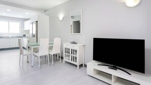 Una sala de estar moderna y bien iluminada con un televisor de pantalla plana, muebles blancos y un comedor con sillas y una mesa.