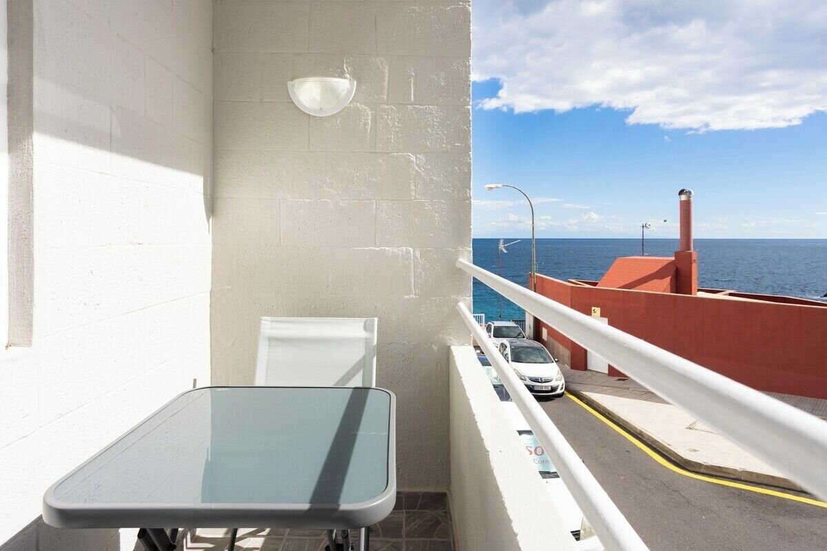 Un balcon avec une table en verre, une chaise blanche et une vue sur la mer et les voitures garées.