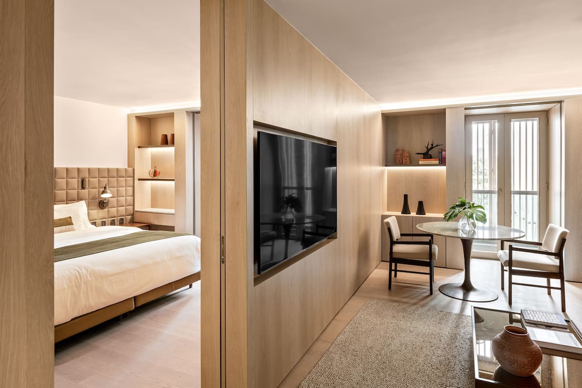 A modern hotel room with a bed, a TV, a dining area, and a large window.