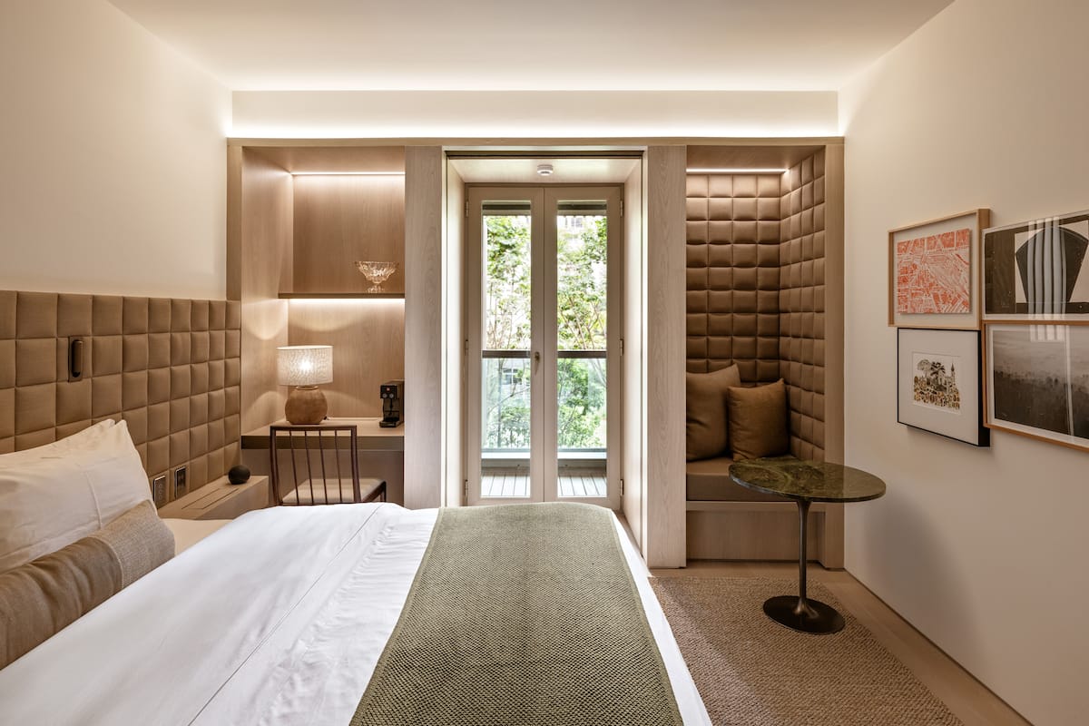 A modern hotel room with a large bed, a seating area with a cushioned bench, a small table, and a view through a glass door.
