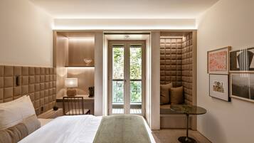 Una habitación de hotel moderna con una cama grande, una zona de estar con un banco acolchado, una mesita y vistas a través de una puerta de cristal.