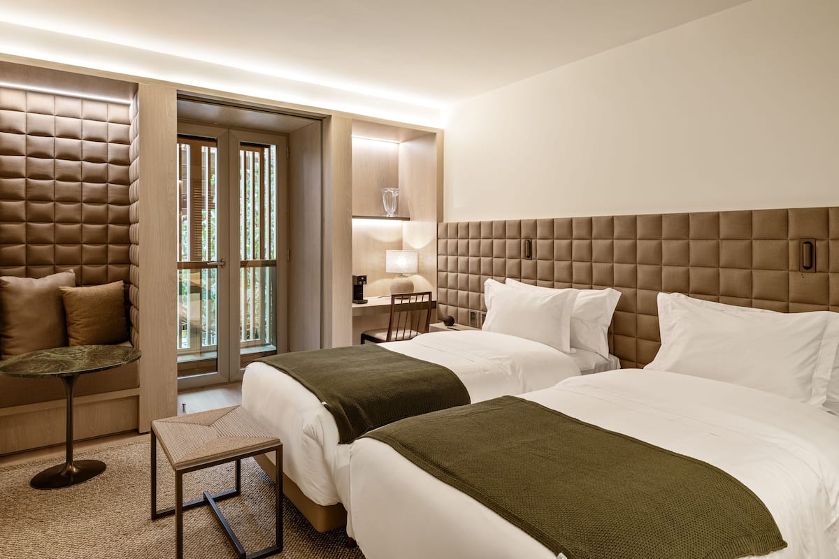 A hotel room with two beds, a sitting area with a chair and table, a lamp, and a view through a door.