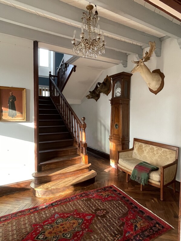 Una escalera con barandillas de madera, un candelabro, una cabeza de ciervo montada en la pared, un sofá y una alfombra estampada.
