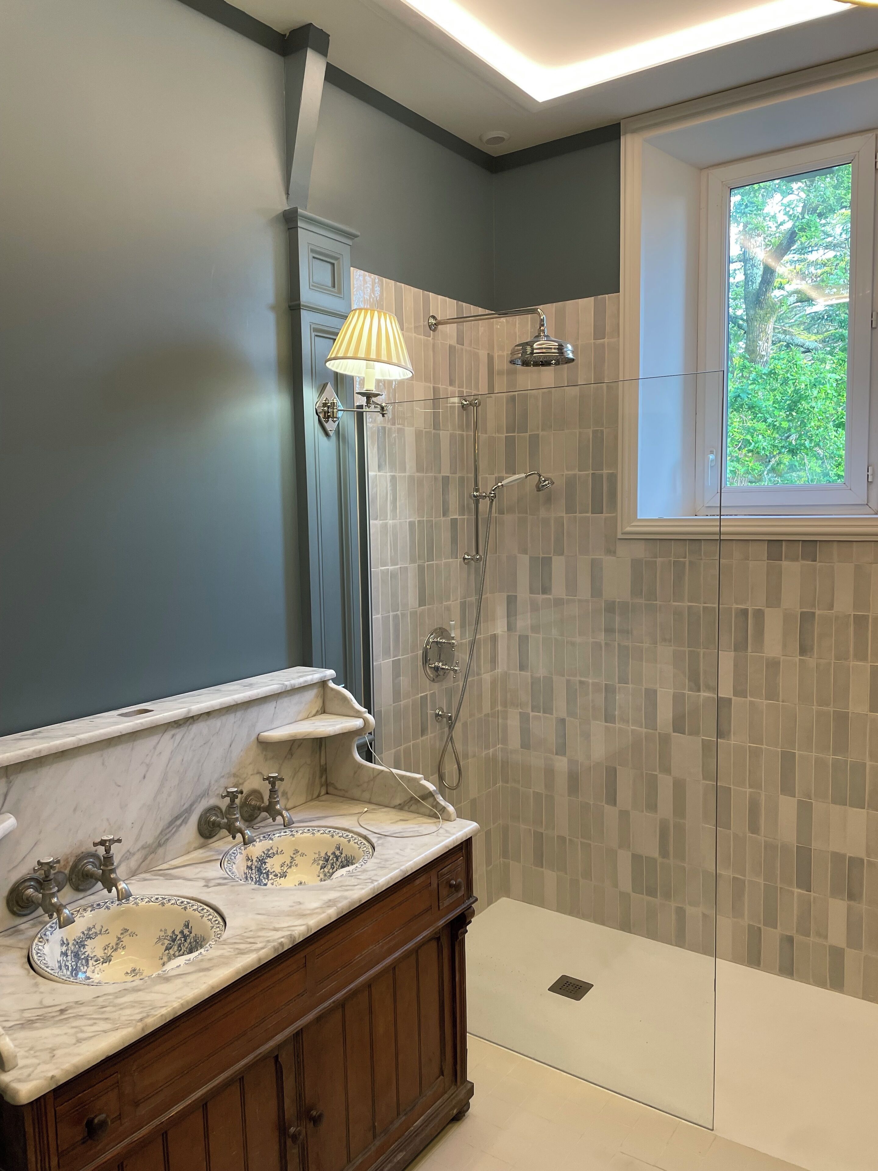 Une salle de bain dotée d’un meuble-lavabo à deux vasques, d’une douche avec une porte en verre et d’une fenêtre laissant entrer la lumière naturelle.