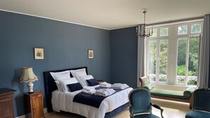 Een slaapkamer met een bed, twee stoelen, een nachtkastje, een lamp en een raam met uitzicht op groen.