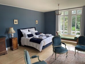 Ein Schlafzimmer mit Bett, zwei Sessel, einem Nachttisch, einer Lampe und einem Fenster mit Blick auf Grünflächen.