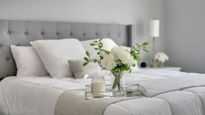 A neatly made bed with white bedding and a vase of white flowers on a bedside table. - Luxurious 3BR, King Bed, Ensuite. Perfect Stay! (Windsor)