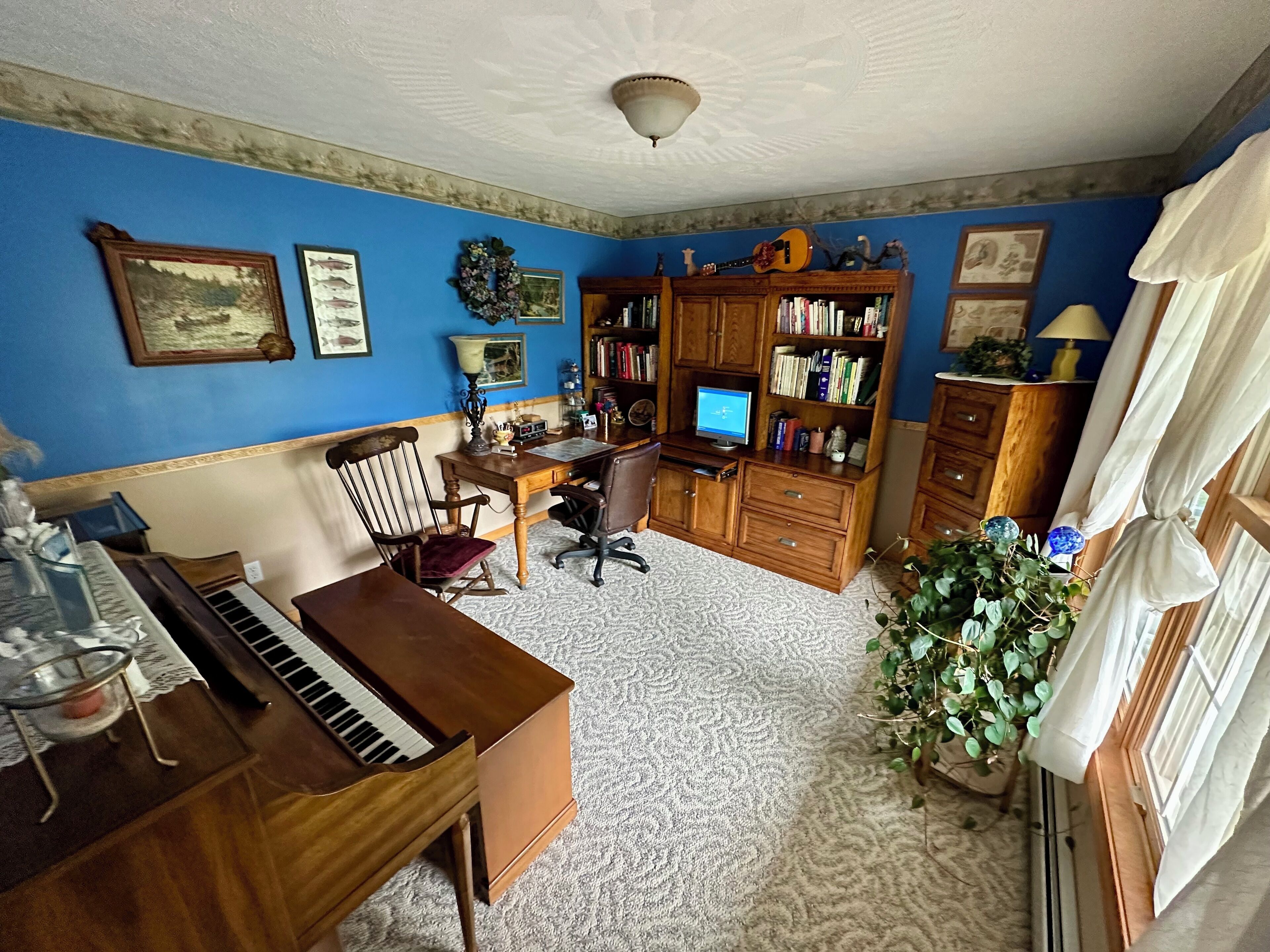 Ein Zimmer mit Klavier, Schreibtisch mit Computer, Bücherregal und Stuhl.