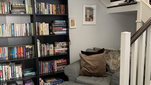 Un soggiorno con un divano grigio, un poggiapiedi grigio, una libreria nera piena di libri, una piccola pianta e un quadro appeso al muro.