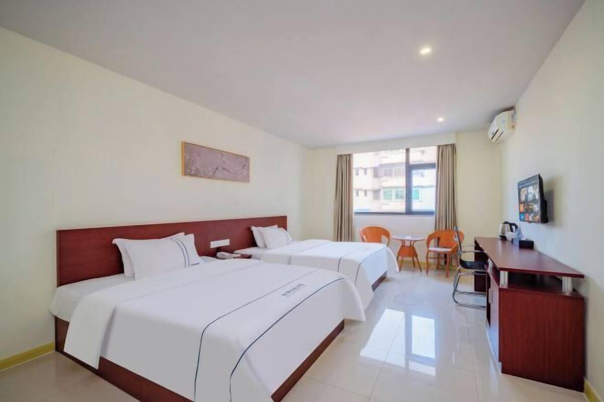 Quarto de hotel com duas camas, uma escrivaninha, cadeiras, uma janela e um quadro na parede.