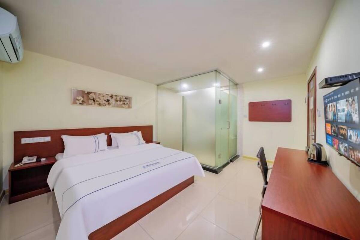 Quarto de hotel com cama, mesa de cabeceira, escrivaninha, cadeira, chuveiro em box de vidro e televisão fixada na parede.