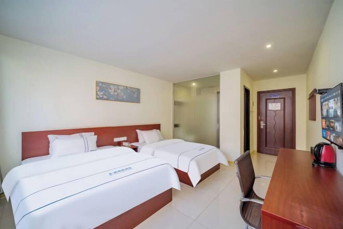 Quarto de hotel com duas camas, uma escrivaninha de madeira, uma cadeira, um quadro na parede e uma porta com um aviso.