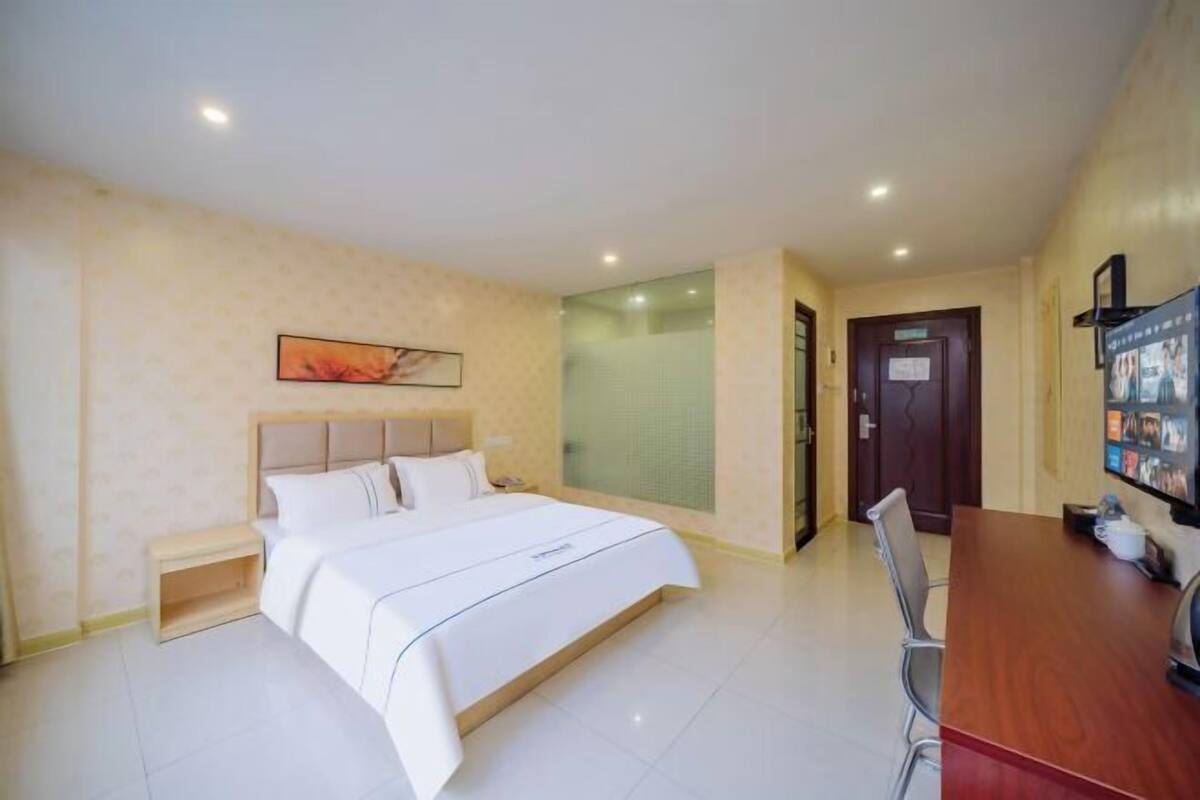Quarto de hotel com cama, mesa de cabeceira, uma cadeira, escrivaninha de madeira e televisão.
