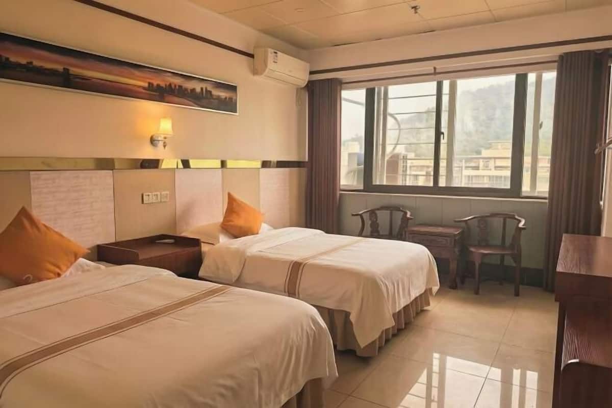 A hotel room with two beds, a desk, a chair, a window with a view, and a painting on the wall.