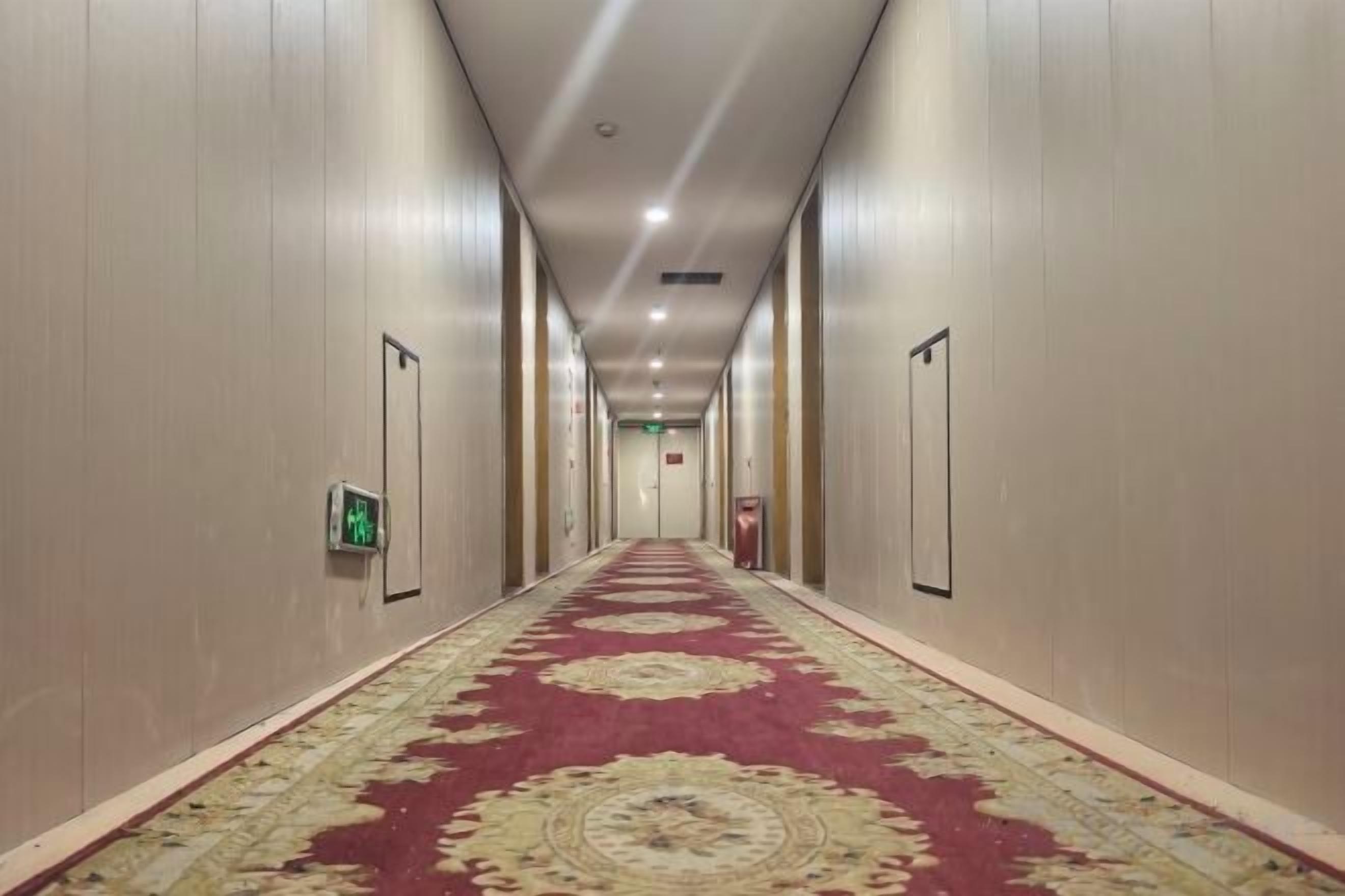 Un pasillo de hotel con alfombra estampada, puertas a ambos lados y un letrero de salida de emergencia.