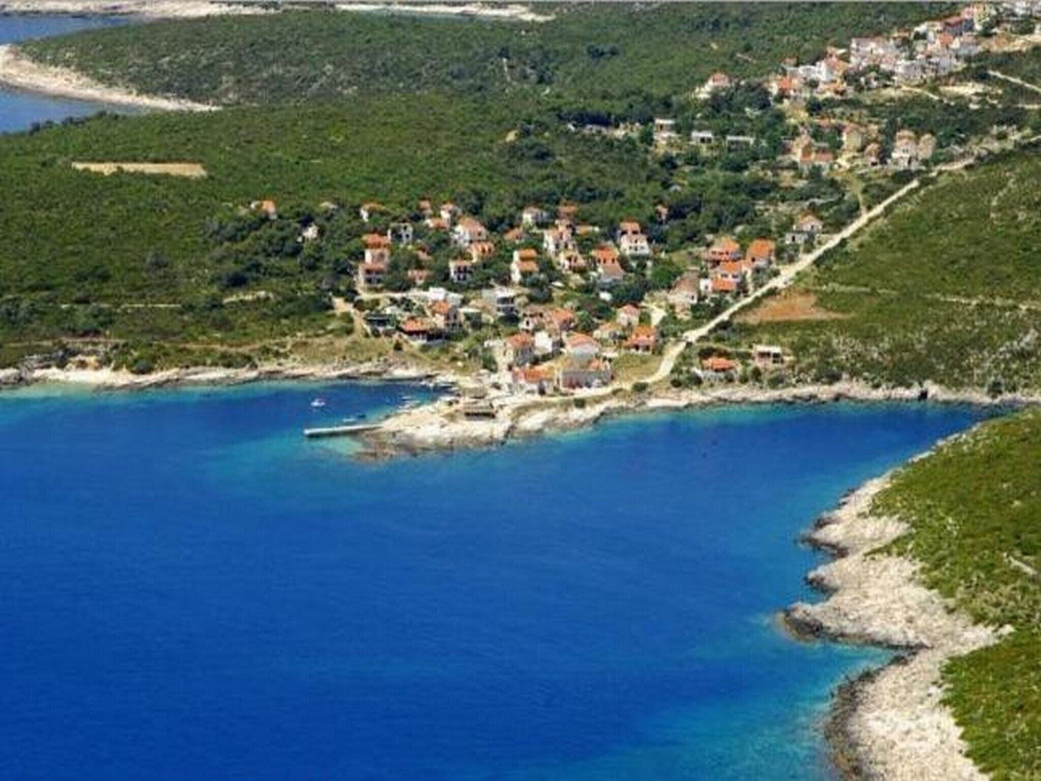 A coastal village with houses along the shoreline, a clear blue sea, and a hilly backdrop.