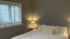 Une chambre avec un grand lit, un ventilateur de plafond, une table de chevet avec une lampe, une fenêtre avec des stores et un panier en osier contenant une serviette.