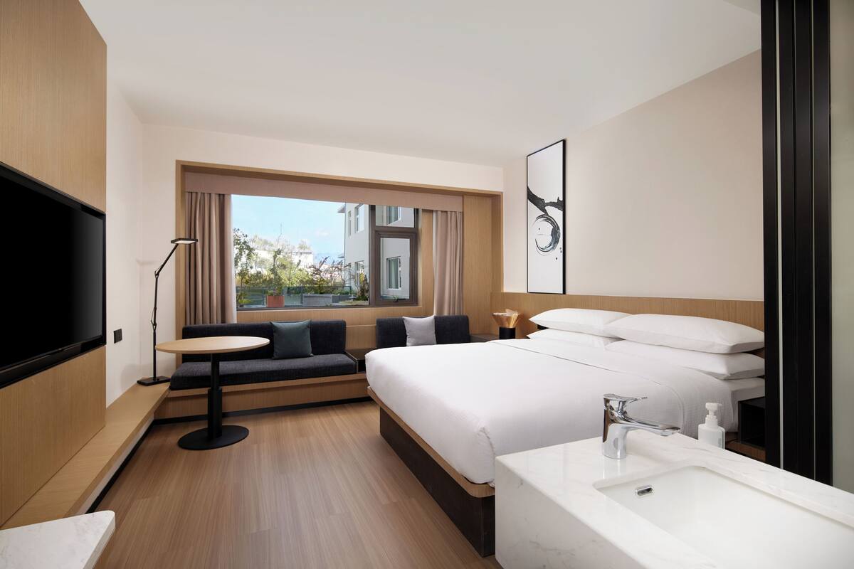 Une chambre d’hôtel moderne avec un grand lit, un canapé, une table ronde, une télévision, une fenêtre avec des rideaux et une salle de bain équipée d’un lavabo et d’un miroir.