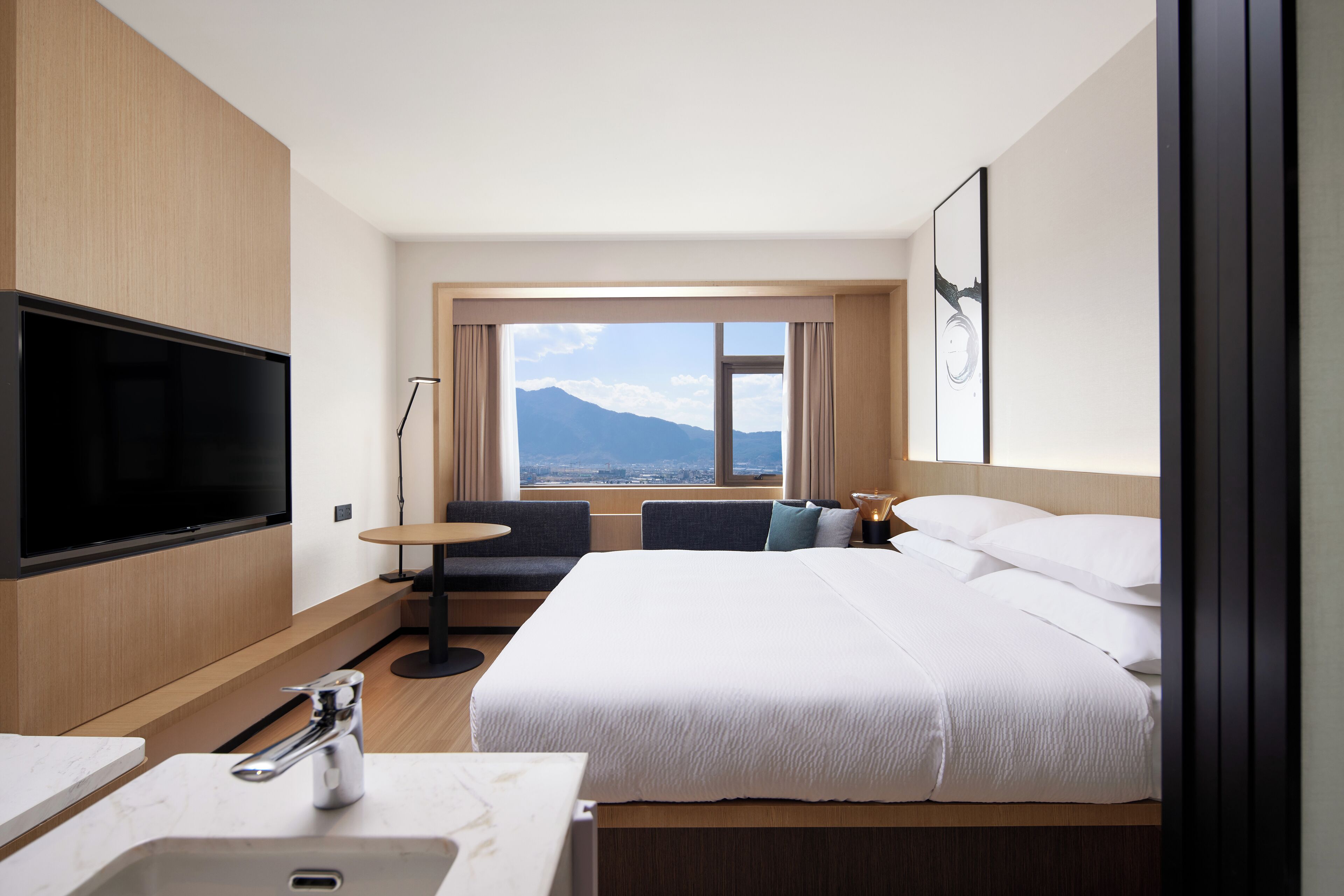 A hotel room with a large bed, a flat-screen TV mounted on the wall, a small round table, and a view of a mountain through the window.