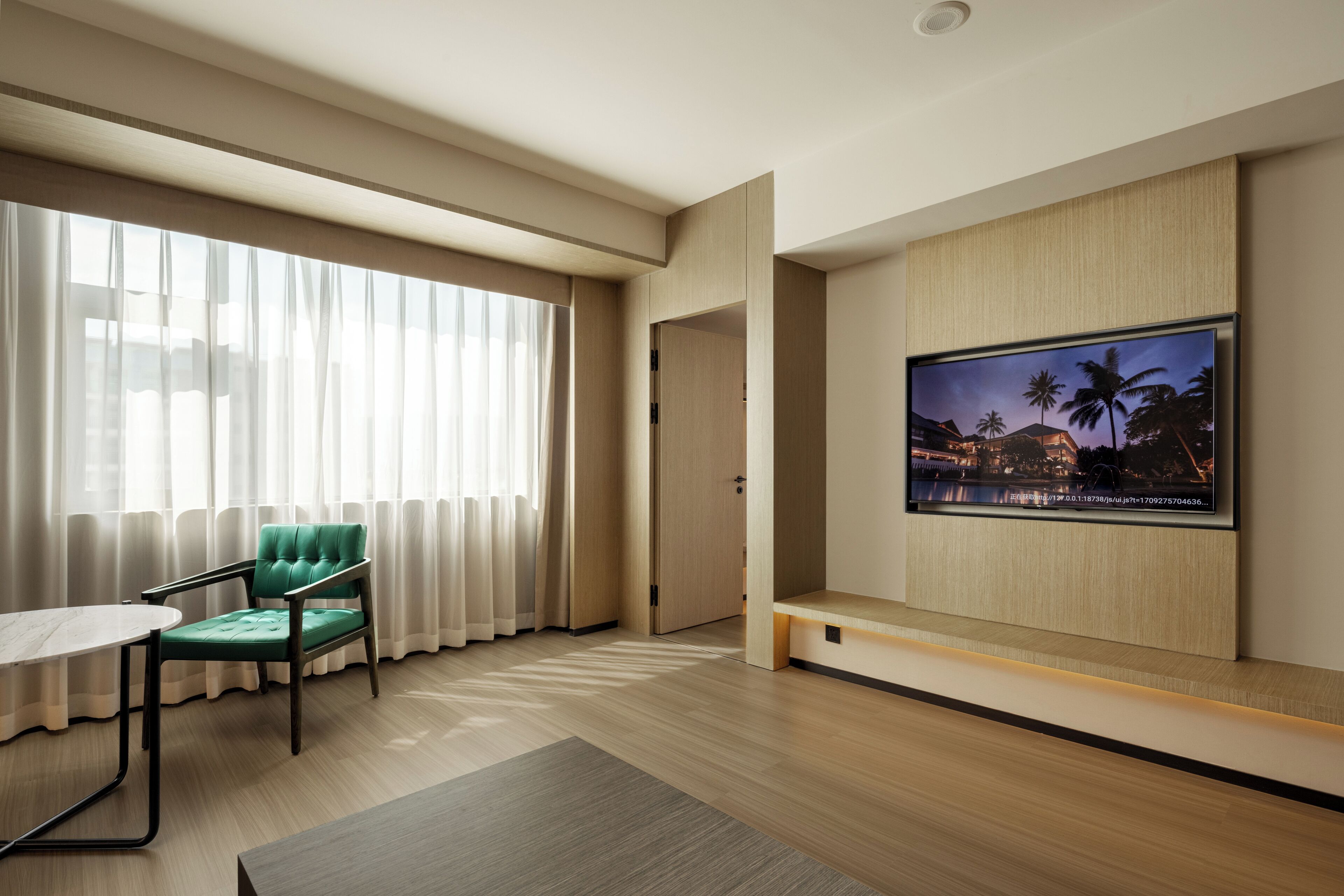 Una habitación de hotel moderna con un ventanal grande, un sillón tapizado en verde, un televisor de pantalla plana montado en la pared y suelo de madera.