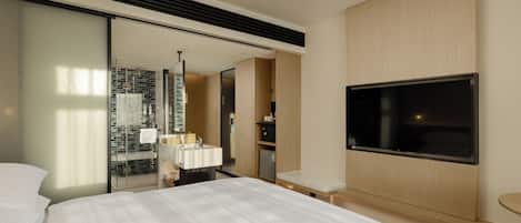 A modern hotel room with a large bed, a flat-screen TV mounted on the wall, a glass shower enclosure, and a wooden vanity with a mirror.