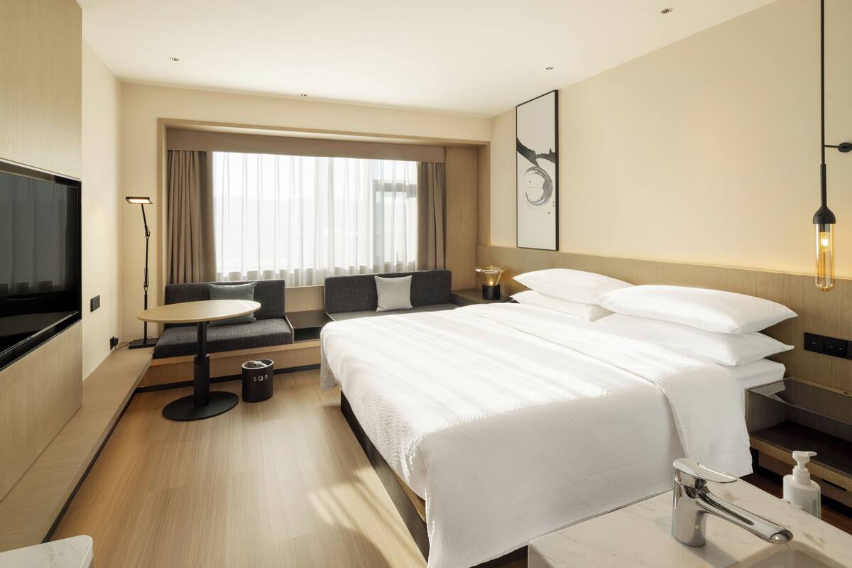 Une chambre d’hôtel comprenant un lit, des tables de chevet, un bureau, une chaise, une télévision, une fenêtre avec des rideaux et une œuvre d’art accrochée au mur.