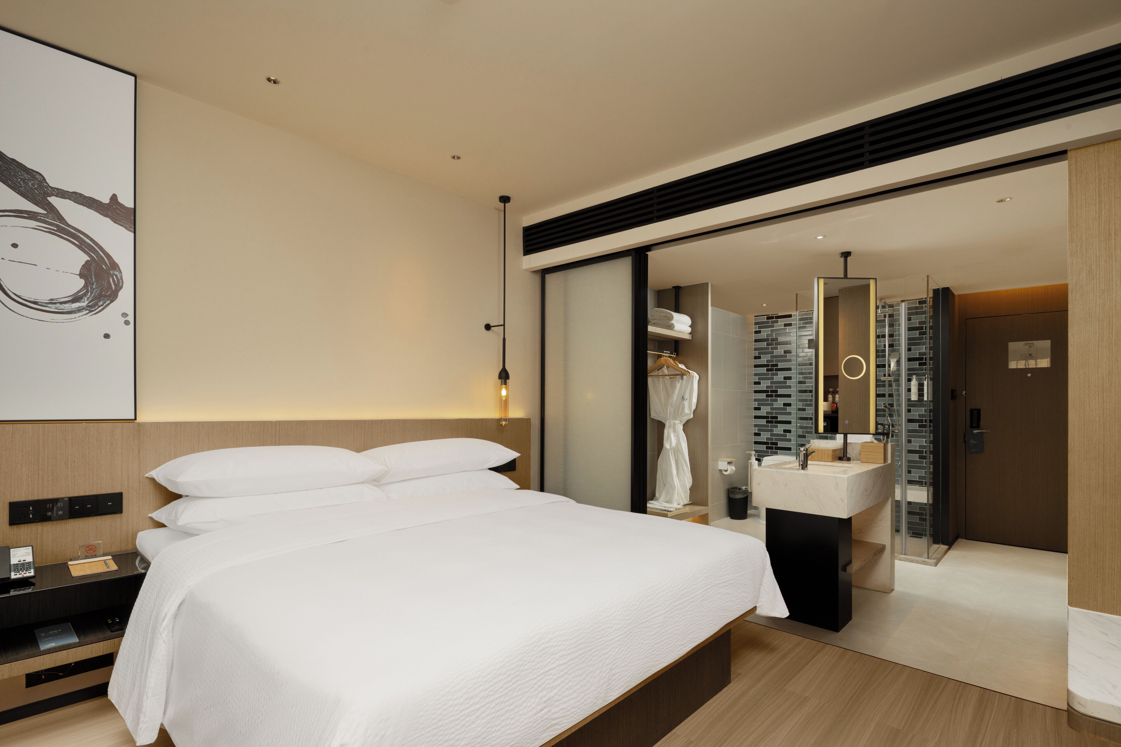A modern hotel room with a large bed, a bedside table with a phone, a mirror, and a bathroom with a sink and glass shower.