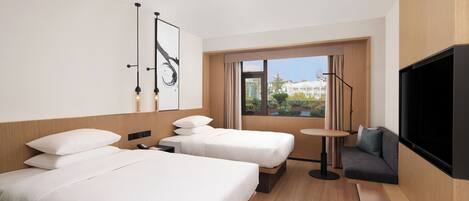 A hotel room with two beds, a sofa, a TV, and a window with a view of buildings and greenery.