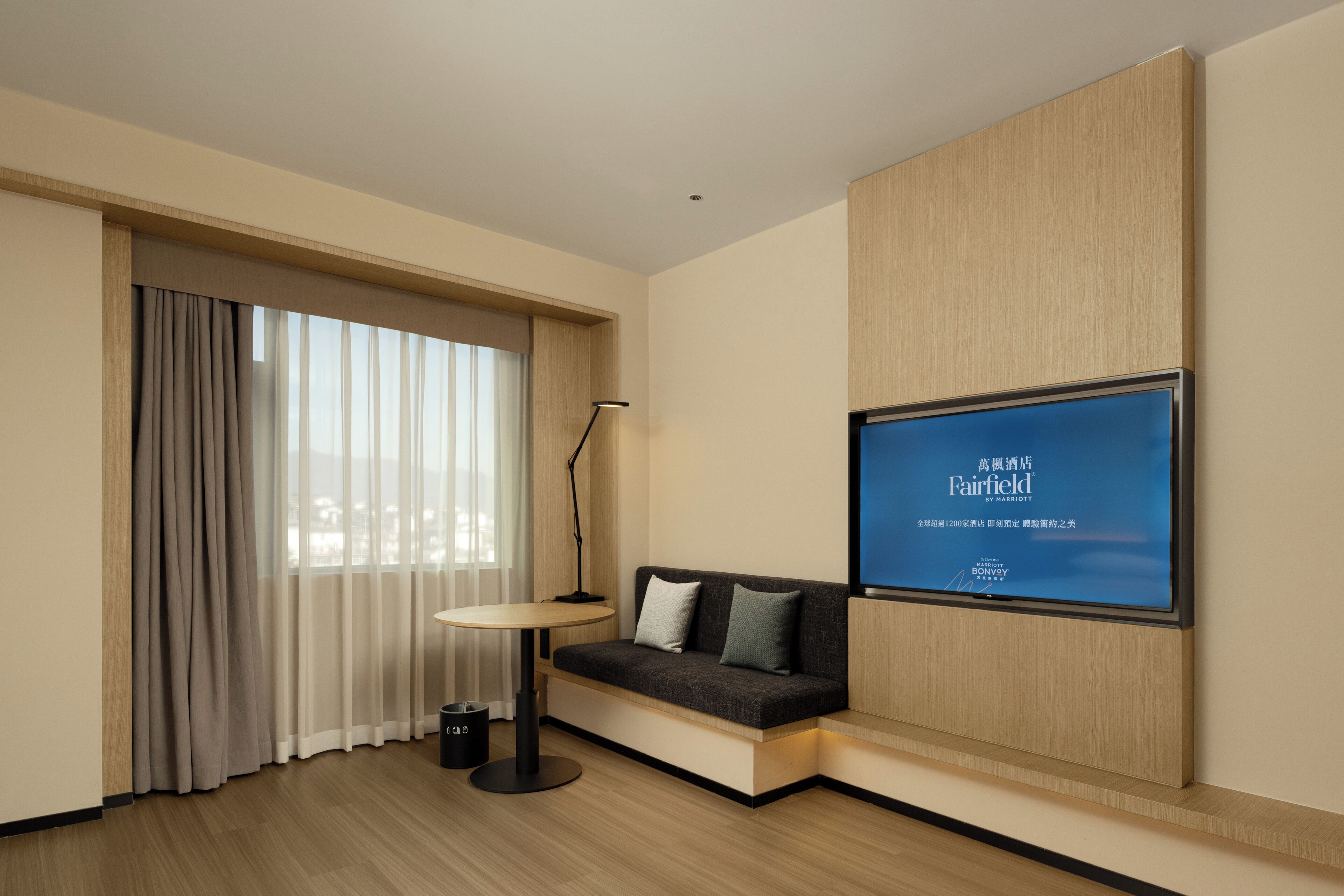 A modern hotel room with a sofa, a small table, a lamp, and a large TV screen displaying "Fairfield by Marriott".