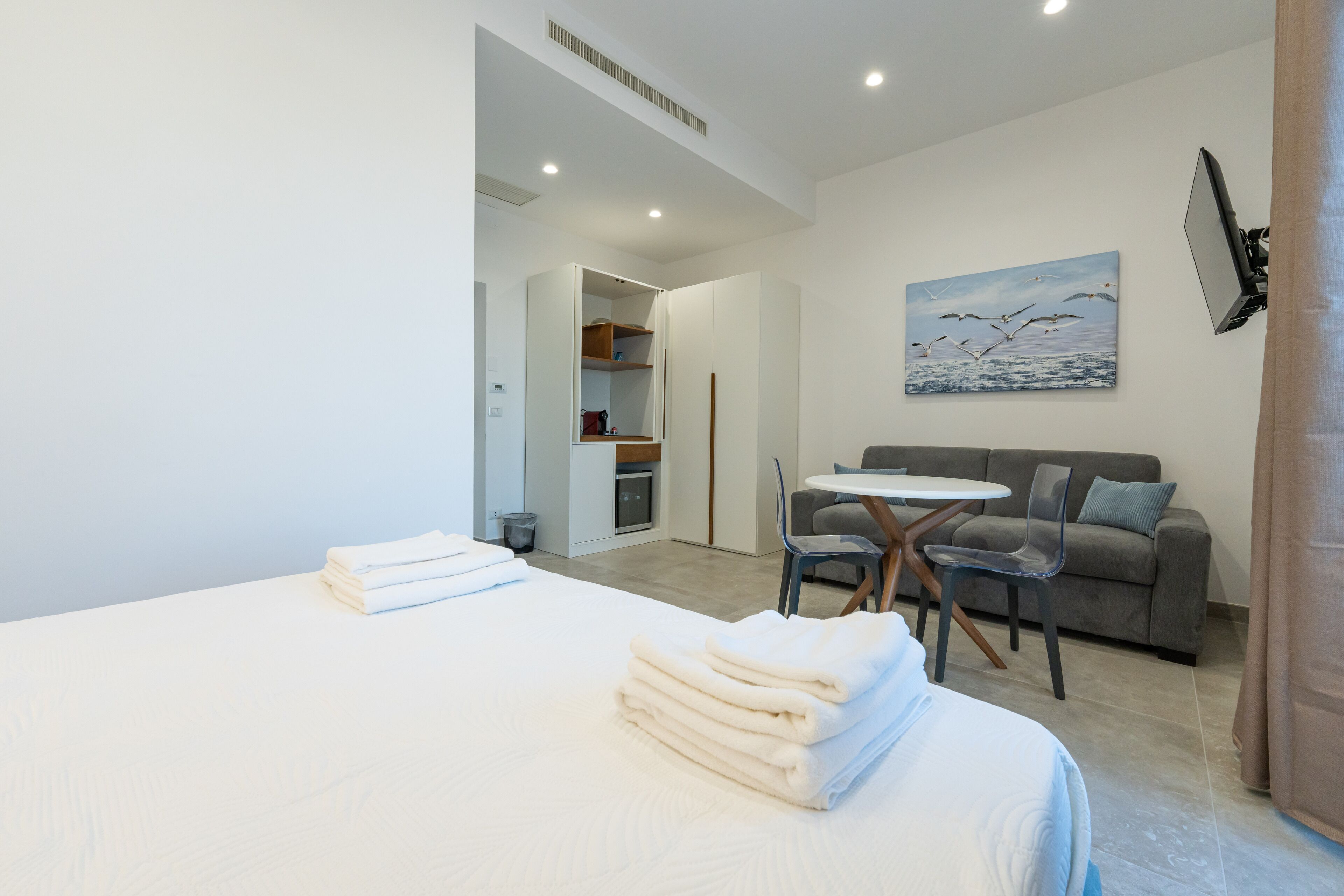 A modern hotel room with a neatly made bed, a dining area with a table and chairs, a sofa, and a painting on the wall.