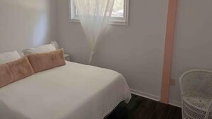 A bedroom with a bed, a ceiling fan, a window with sheer curtains, and a white chair.