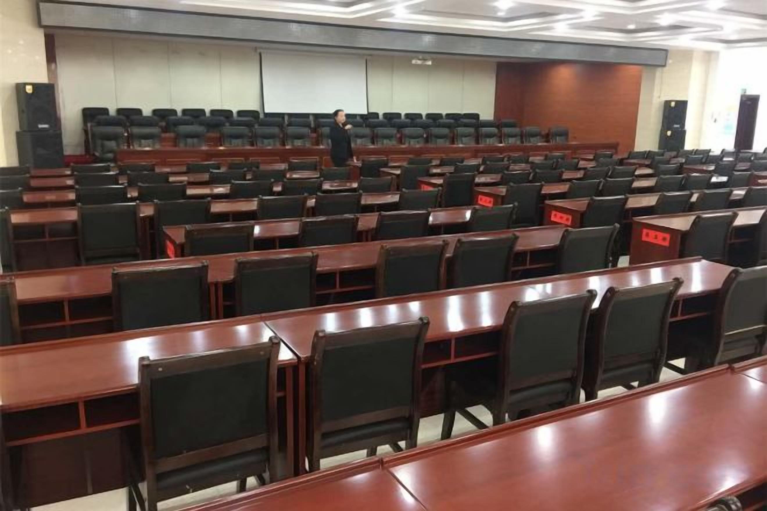 A conference room with rows of tables and chairs, a podium, and a person standing at the front.