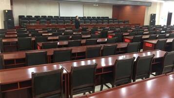 A conference room with rows of tables and chairs, a podium, and a person standing at the front.