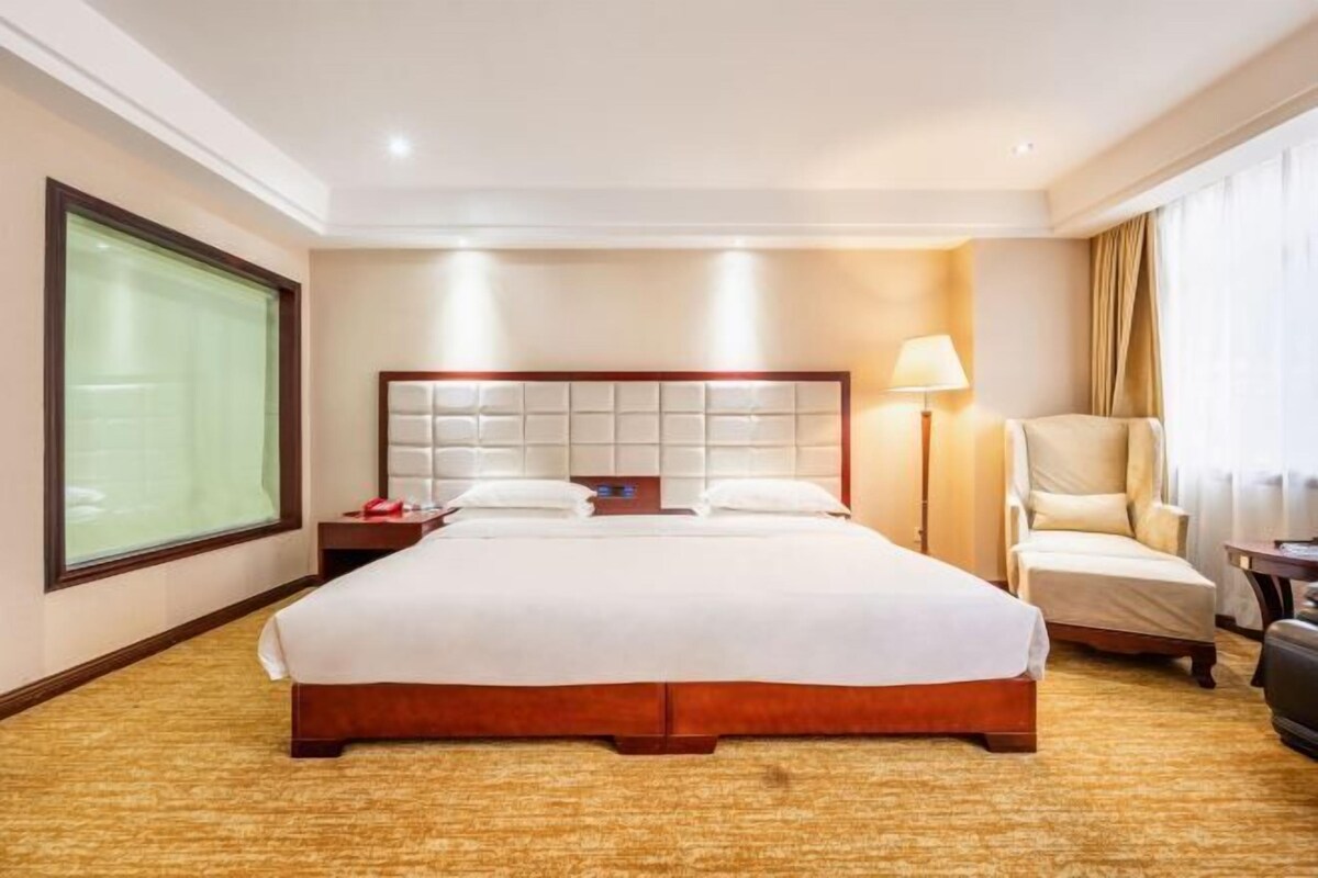 Habitación de hotel con una cama grande, una silla, una mesita de noche, una lámpara y vistas al exterior.