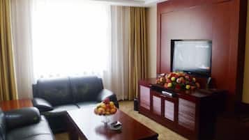 A living room with a leather sofa, a wooden coffee table with fruit, a television on a cabinet, and a large window with curtains.