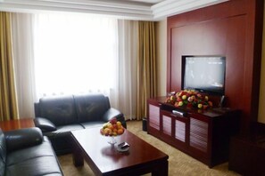 Un salón con un sofá de cuero, una mesa de centro de madera con fruta, un televisor sobre un mueble bajo y un ventanal con cortinas.
