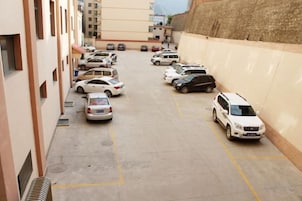 Un estacionamiento con varios coches aparcados en filas, junto a un edificio con un muro alto.